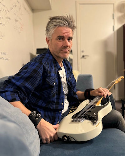 Man with grey hair and blue plaid shirt holds a white guitar on a blue couch in a room with a white door and wall covered in signatures.