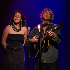 Two musicians perform on stage, one singing and one playing guitar, under purple and blue lights. They stand before vintage microphone and a light.