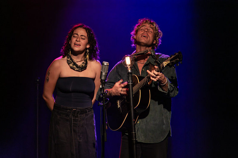 Two musicians perform on stage, one singing and one playing guitar, under purple and blue lights. They stand before vintage microphone and a light.
