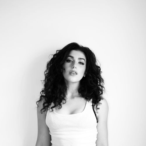 Woman with curly hair (Caroline Romano) in a white tank top stands against a plain white background, displaying a calm and confident expression. Black and white photo.