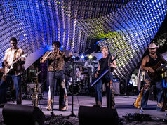The Free Label blue-lit geometric canopy. Five musicians with instruments, lively atmosphere, and vibrant clothing patterns.