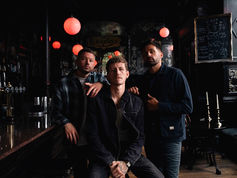 Three men in a dimly lit bar with red lamps, one seated, two standing. Chalkboard menu visible. Moody atmosphere.