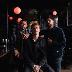 Three men in a dimly lit bar with red lamps, one seated, two standing. Chalkboard menu visible. Moody atmosphere.