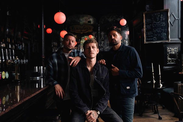 Three men in a dimly lit bar with red lamps, one seated, two standing. Chalkboard menu visible. Moody atmosphere.
