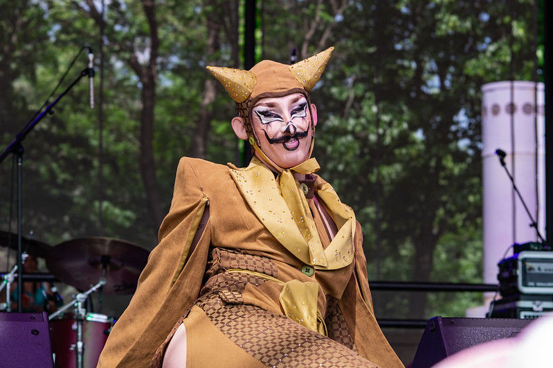 Drag performer in gold horned costume and dramatic makeup poses on an outdoor stage, with trees and microphones behind.