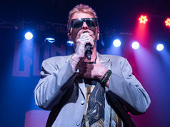 Singer in sunglasses performs onstage with microphone under blue and pink lights, hand on chest, at a live concert.