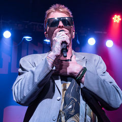 Singer in sunglasses performs onstage with microphone under blue and pink lights, hand on chest, at a live concert.