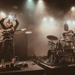 Two polka-dot-clad musicians perform on a smoky stage, one raising a guitar overhead while another plays drums under bright lights.