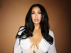 Woman with long black hair wearing a white top, standing against a solid brown background, looking confidently at the camera.
