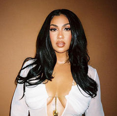 Woman with long black hair wearing a white top, standing against a solid brown background, looking confidently at the camera.