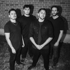 Four men pose in black-and-white before a brick wall at night, standing on grass with serious expressions.