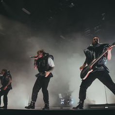 Three performers in dark puffy outfits sing and play guitar energetically on a fog-filled stage, illuminated by dramatic lighting.