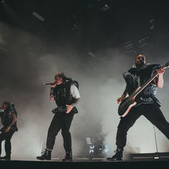 Three performers in dark puffy outfits sing and play guitar energetically on a fog-filled stage, illuminated by dramatic lighting.