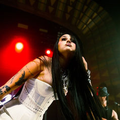 Performer with long black hair and tattoos sings passionately on stage, wearing a white dress. Red lights glow in background, creating a dramatic mood.