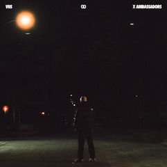 A person stands alone on a dark street under a streetlight. Text reads "VHS" and "X Ambassadors." The mood is mysterious and solitary.