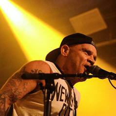 Musician wearing a black cap sings into a microphone on stage, bathed in yellow light. Tattoos visible on arms. Shirt reads "NEW".