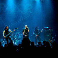 Band performs energetically on stage under blue lights. Crowd is visible in the foreground. "RAZOR" logo is displayed on the backdrop.