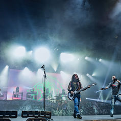 Rock band performs on a smoky stage with bright blue-white lights and a huge screen behind them.