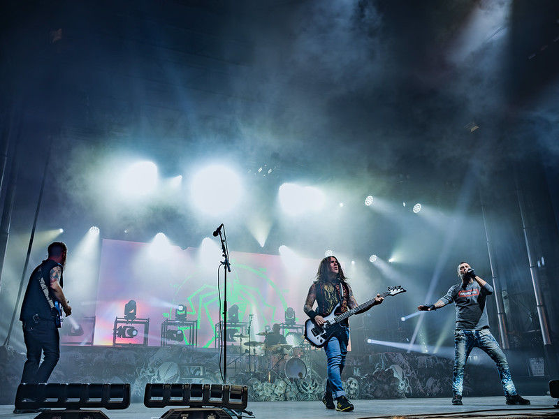 Rock band performs on a smoky stage with bright blue-white lights and a huge screen behind them.