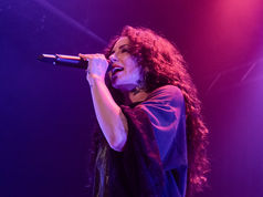Singer with curly hair performs passionately under purple and pink stage lights, holding a microphone. The mood is energetic and vibrant.