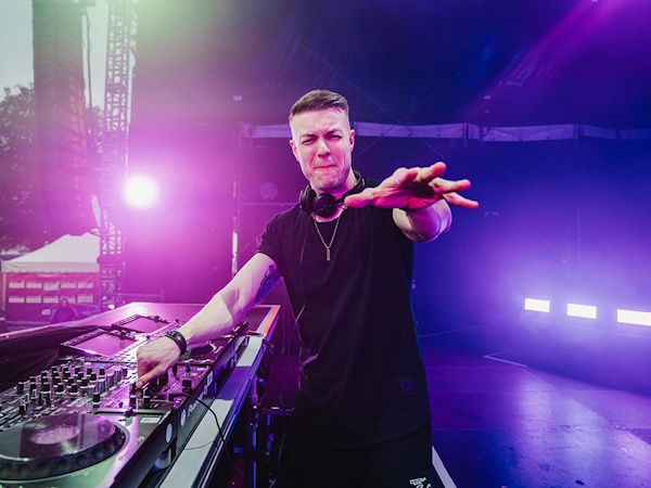 DJ in a black shirt performs at a mixer on a purple-lit stage, reaching toward the crowd.