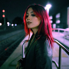 Woman with red hair stands on a dimly lit train platform at night, wearing a dark jacket. A moody, urban ambiance surrounds her.