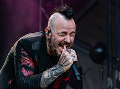 Tattooed singer Adam Gontier with a mohawk belts into a microphone onstage under dark purple lighting, eyes closed in intensity