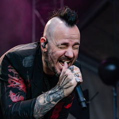 Tattooed singer Adam Gontier with a mohawk belts into a microphone onstage under dark purple lighting, eyes closed in intensity