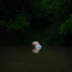 Person crouches half-submerged in dark forest water, surrounded by dense green foliage.