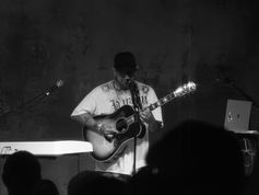 Black-and-white photo of a guitarist singing into a microphone on stage, wearing a cap, with a laptop and audience silhouettes.