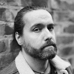 Man with a beard leans against a brick wall, looking intently at the camera. Black-and-white image, casual jacket with a fur collar.