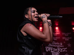 Female singer with braids belts into a microphone on a red-lit stage, before an Off the Rez Tour backdrop.