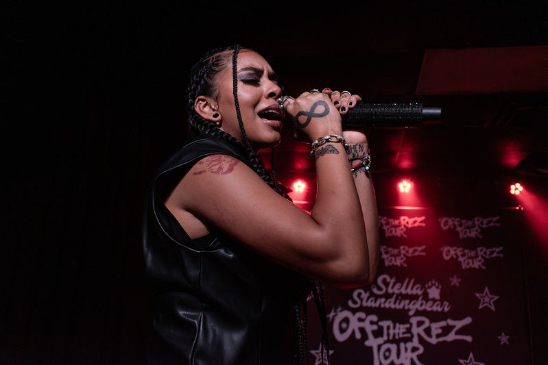 Female singer with braids belts into a microphone on a red-lit stage, before an Off the Rez Tour backdrop.