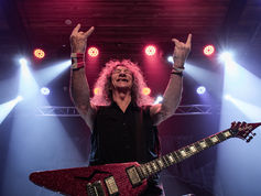 Smiling rock guitarist with a glittery red electric guitar raises both hands throwing up devil horns on a lit stage with pink, red, and blue spotlights.