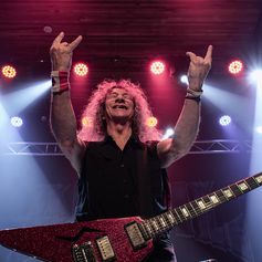 Smiling rock guitarist with a glittery red electric guitar raises both hands throwing up devil horns on a lit stage with pink, red, and blue spotlights.