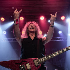 Smiling rock guitarist with a glittery red electric guitar raises both hands throwing up devil horns on a lit stage with pink, red,  and blue spotlights.