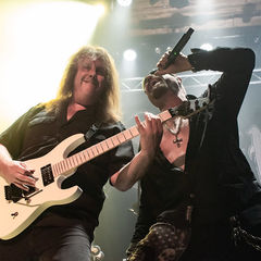 Two musicians on stage: one plays a white electric guitar, the other sings into a mic. Intense lighting, drum set in background, energetic mood.