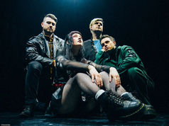 Four people pose on stage in dark, edgy clothing. The mood is intense. A woman sits in front, and three men stand behind in a dimly lit scene.