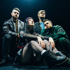 Four people pose on stage in dark, edgy clothing. The mood is intense. A woman sits in front, and three men stand behind in a dimly lit scene.
