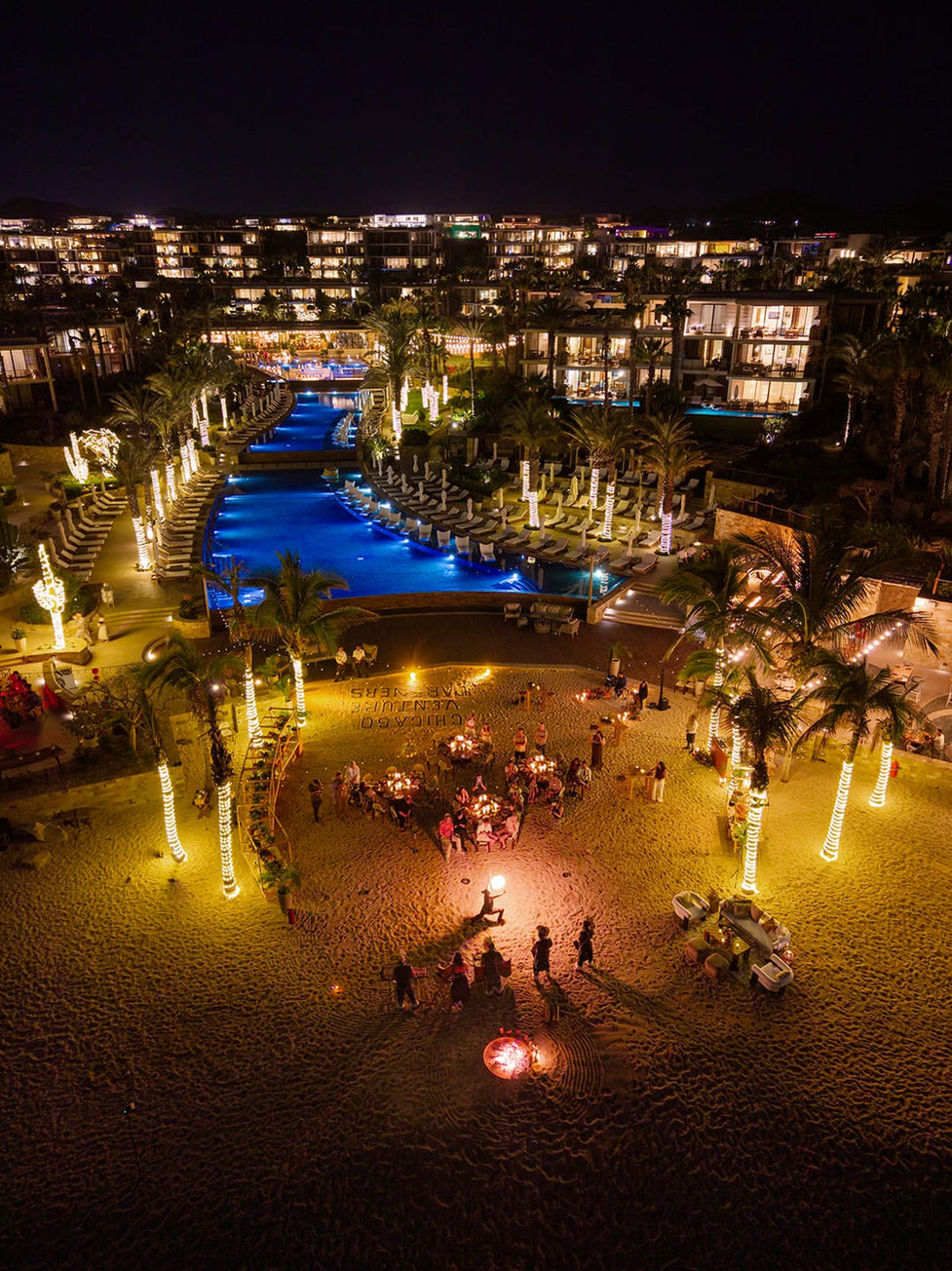 Wide aerial nighttime view of Chileno Bay Resort in Los Cabos during a wedding event, with the resort's illuminated infinity pool glowing blue, palm trees wrapped in warm lights, a candlelit beach celebration below, and the resort's modern terraced building lit up behind.