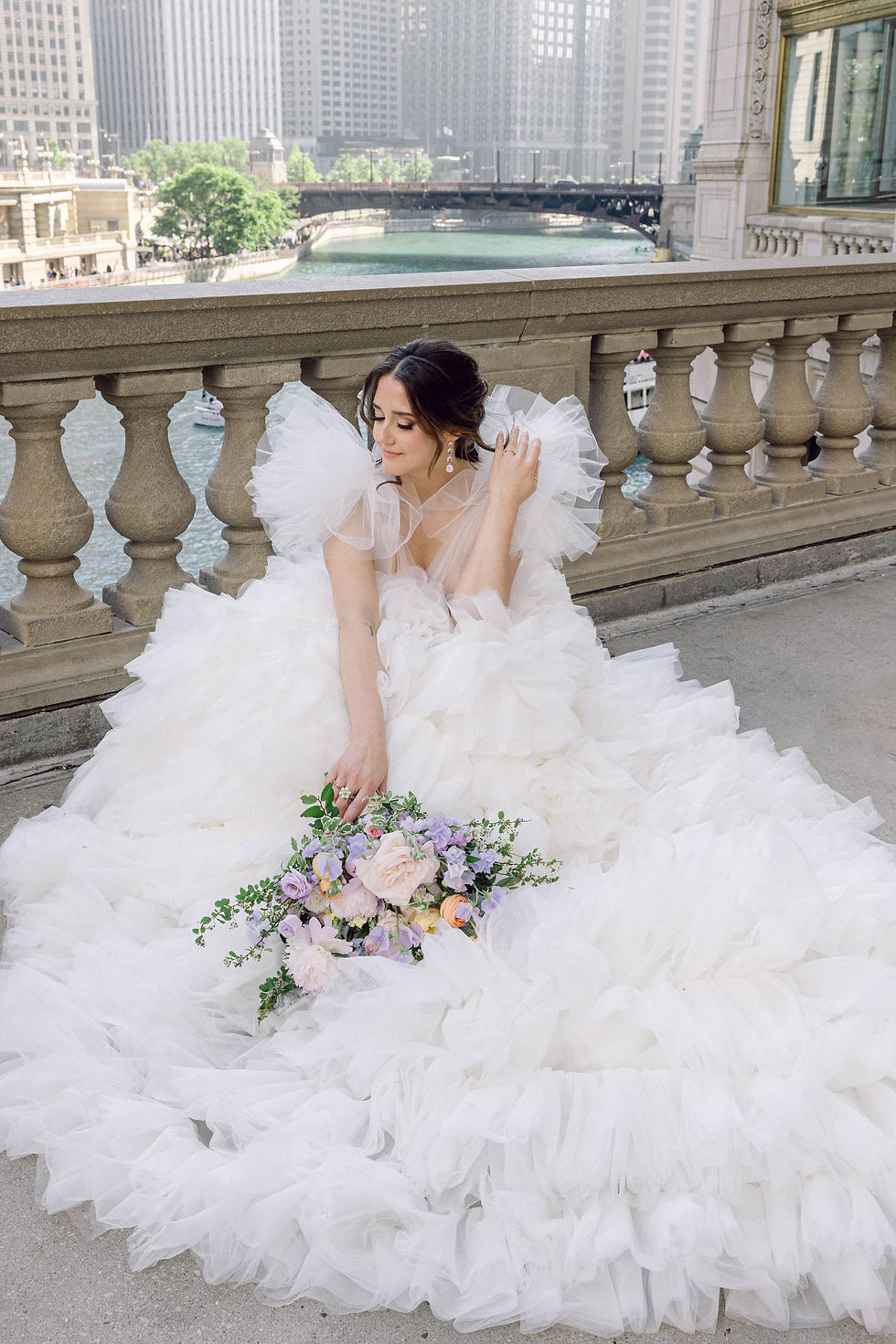 Bride seated along the Chicago River in a dramatic ruffled tulle gown holding a pastel bouquet, showcasing couture bridal fashion.