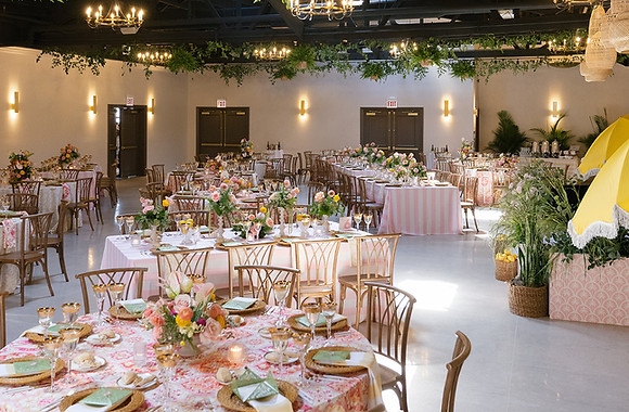 Elegant wedding reception setup at Bloom Events in Chicago featuring floral-patterned linens, pastel florals, natural wood chairs, and bright yellow umbrellas beneath greenery-draped chandeliers.