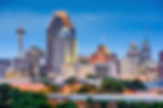 San Antonio skyline at dusk featuring the illuminated Tower Life Building, the Tower of the Americas, and other downtown skyscrapers against a blue evening sky.