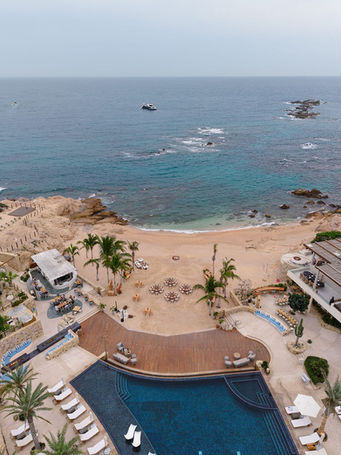 Aerial daytime view of Chileno Bay Resort in Los Cabos showing the protected cove, white sand beach set with wedding reception tables, the wooden event deck, pool terrace, and the deep turquoise Sea of Cortez with rocky outcroppings.