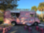 A pink food truck labeled "Las Mexicanas" is parked outdoors with pink stools and tables set up in front, surrounded by palm trees and greenery.