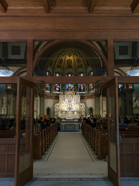Interior view of three towering stained glass windows at St. Vincent de Paul Church in Chicago, depicting biblical scenes in rich jewel tones of blue, red, green, and gold, framed by white stone arches and carved ornamental details.