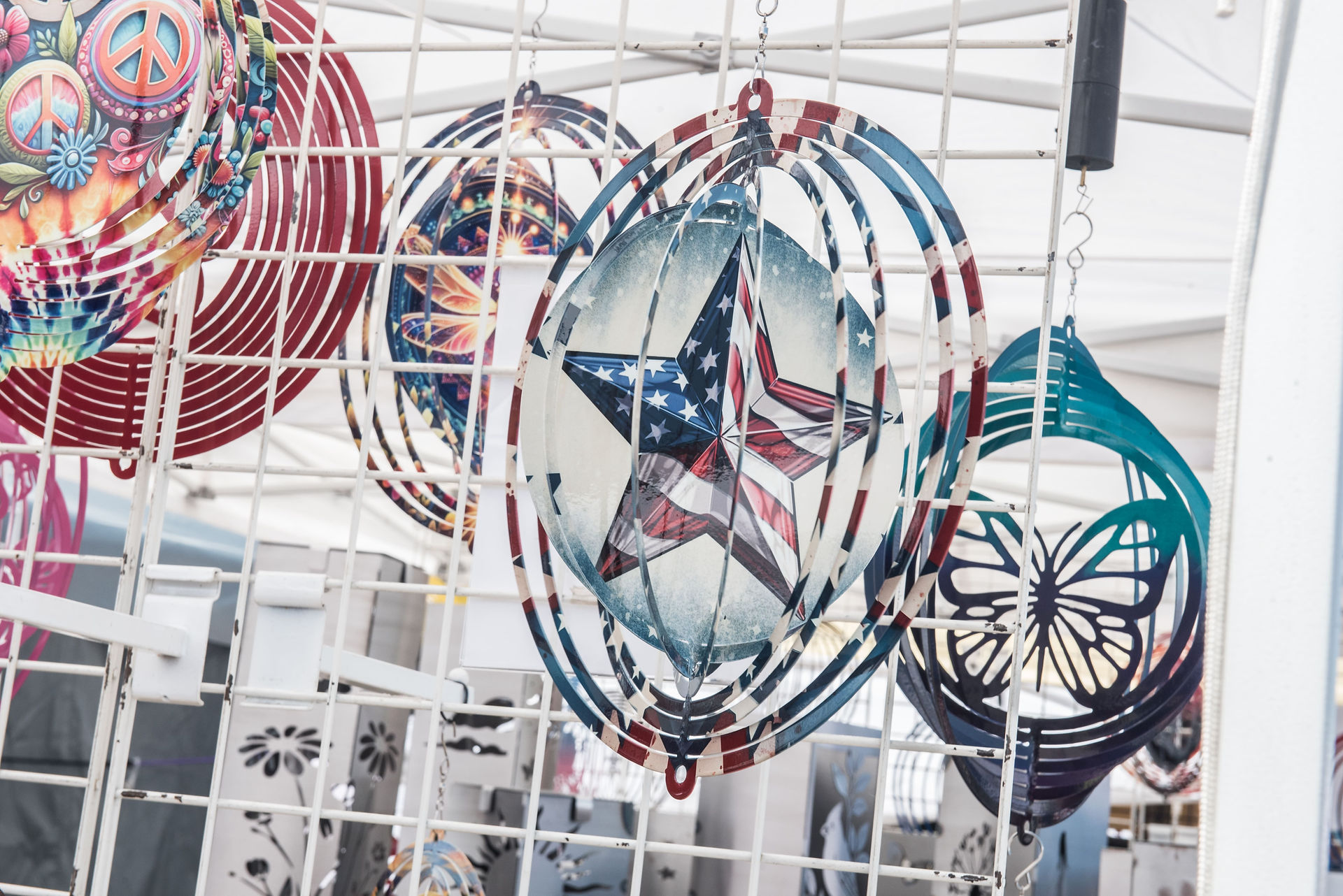Decorative hanging metal wind spinners in patriotic star, peace sign, and butterfly designs on display at an Old Canal Days 2026 artisan market vendor booth