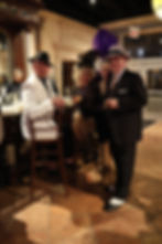 Flapper character in vintage costume poses behind a bar with mobster-themed guests at a speakeasy-style event.