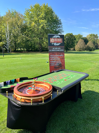 Outdoor casino game setup with a roulette wheel, blackjack table layout, and stacks of chips on a grassy lawn.