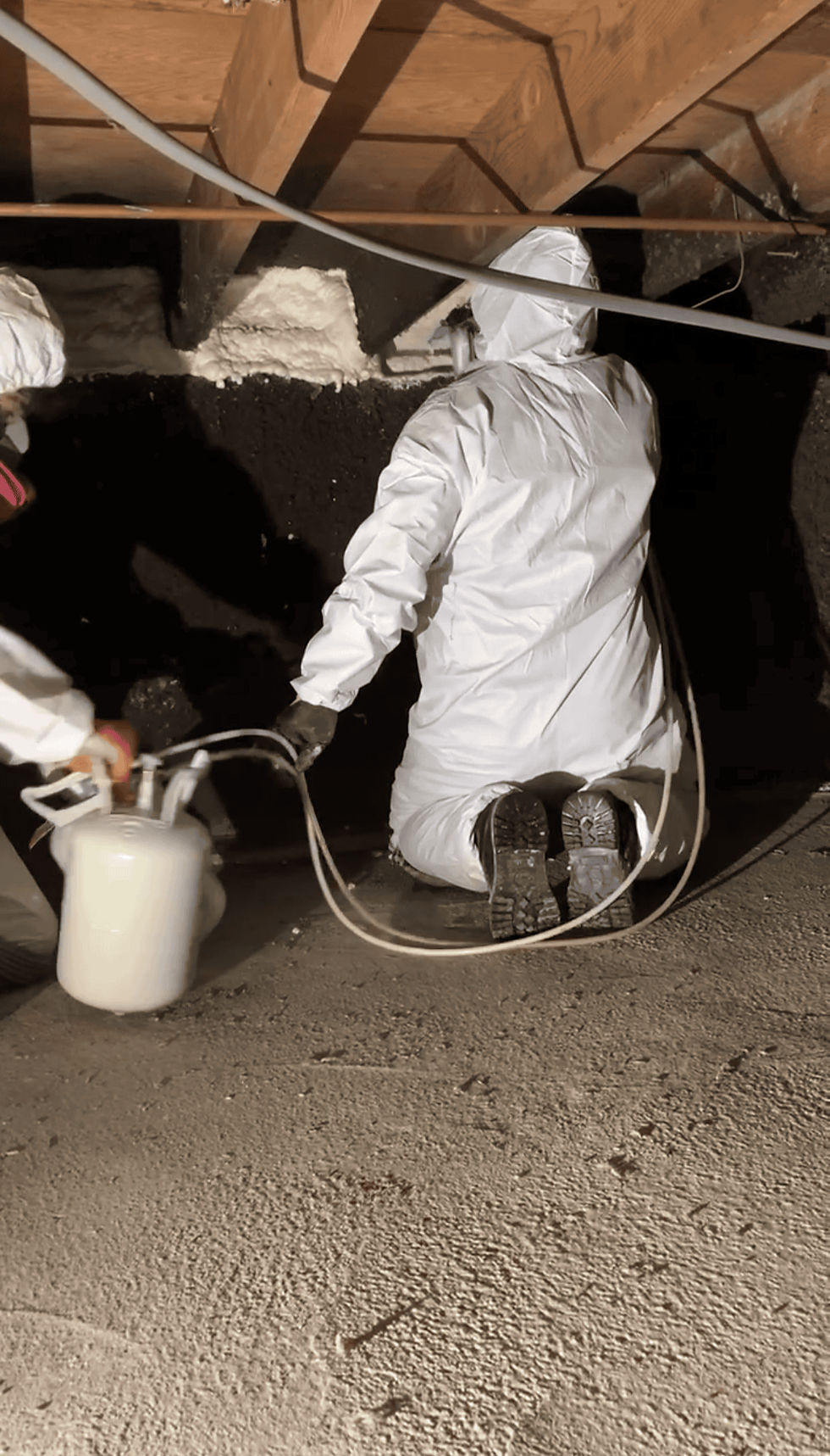 Technician in full protective Tyvek suit applying spray foam insulation to crawl space walls and rim joists during encapsulation